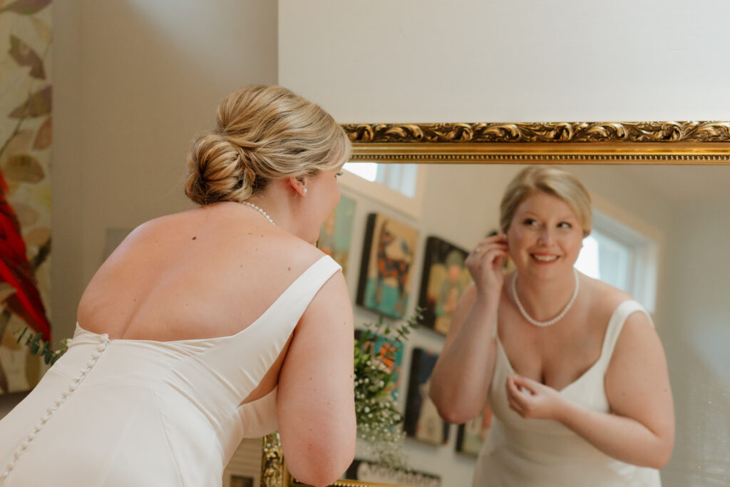 Bride putting on her earrings on her wedding day while looking at her reflection in the mirror.