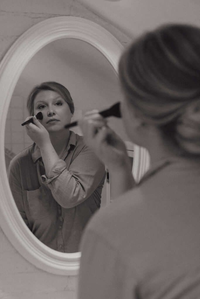 Bride putting on makeup on her wedding day while looking at her reflection in the mirror.