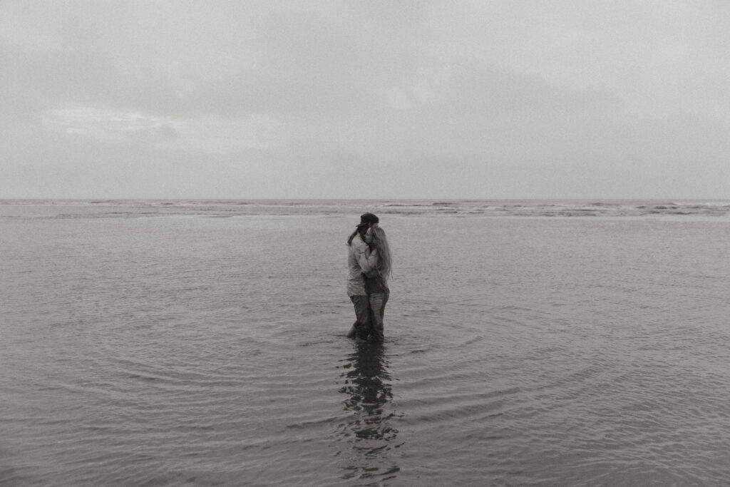 A black and white photo of a couple playing in the ocean during their sunrise engagement session at Folly Beach in South Carolina.